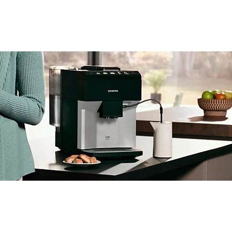 Eine Kaffeemaschine mit Milchaufschäumer. Daneben Gebäck und Obst. Im Hintergrund eine Frau.