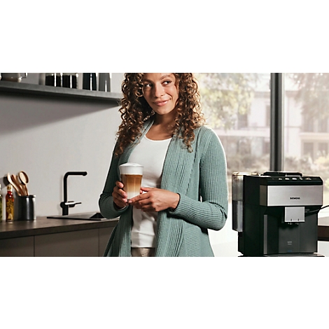 Frau hält einen Latte Macchiato in der Küche, Kaffeemaschine im Hintergrund.