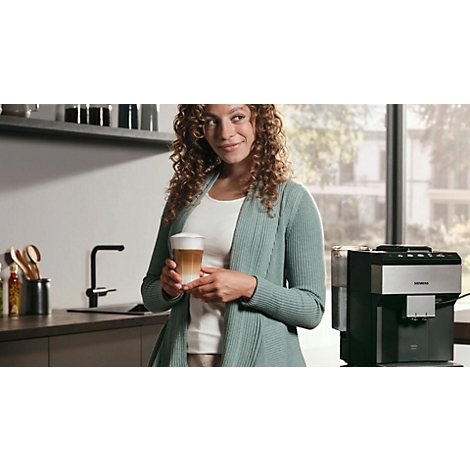 Frau mit lockigem Haar hält Latte Macchiato, lächelnd in der Küche. Kaffeemaschine im Hintergrund.