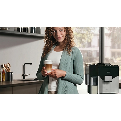 Eine Frau mit lockigem Haar hält einen Latte Macchiato in der Küche. Im Hintergrund eine Kaffeemaschine.