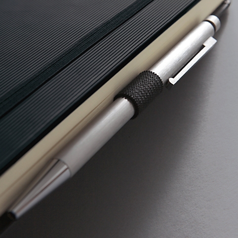 Gros plan : stylo argenté inséré dans un cahier. Des lignes sombres structurent les pages, avec une couverture claire à côté.