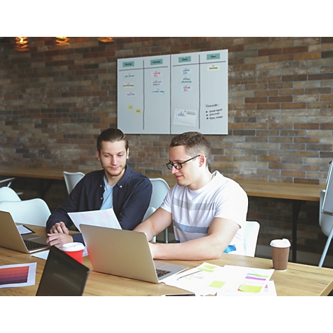 Twee mannen werken aan laptops aan een tafel. Een bakstenen muur en een whiteboard op de achtergrond.