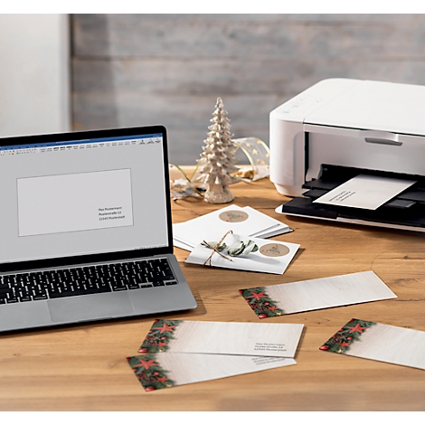 Ordinateur portable et imprimante sur une table en bois. Des cartes de Noël sont imprimées. Sapin de Noël en arrière-plan.