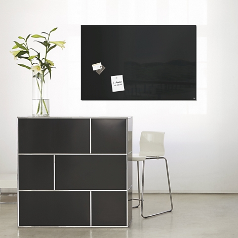 Schwarze Magnettafel an weißer Wand, darunter eine schwarze Kommode mit silbernen Elementen und ein Stuhl.