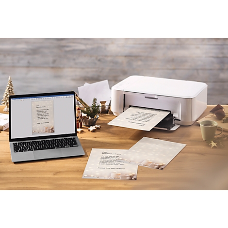 Een laptop en een printer printen kerstkaarten op een houten tafel. Decoraties zoals sterren en een kerstboom zijn op de achtergrond te zien.