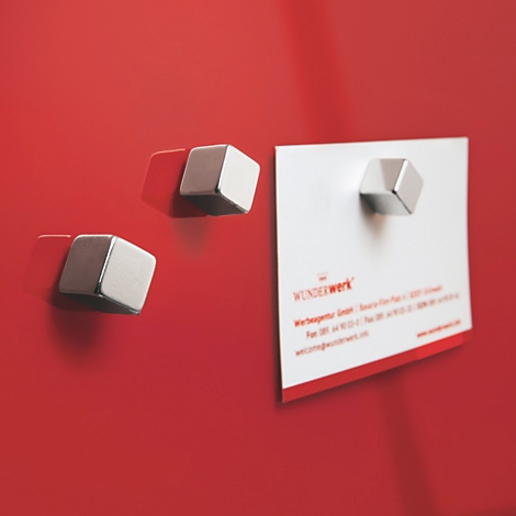 Drei metallische, geometrisch geformte Magnete haften an einer roten Oberfläche, einer hält eine Visitenkarte.