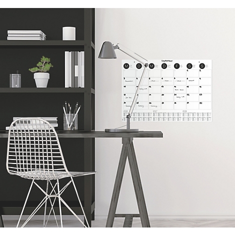 Schreibtisch mit Lampe und Kalender an der Wand, daneben ein Regal und Stuhl.