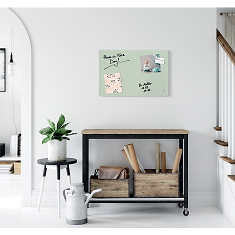 Tableau magnétique vert clair avec des notes et une photo, sur un mur blanc. En dessous, une table avec décor.