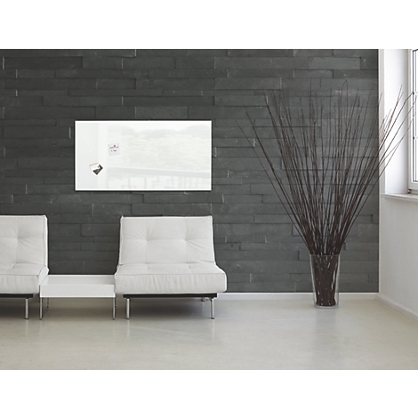 Un tableau magnétique en verre blanc sur un mur de pierre sombre. En dessous, deux fauteuils blancs et un vase avec des graminées ornementales.