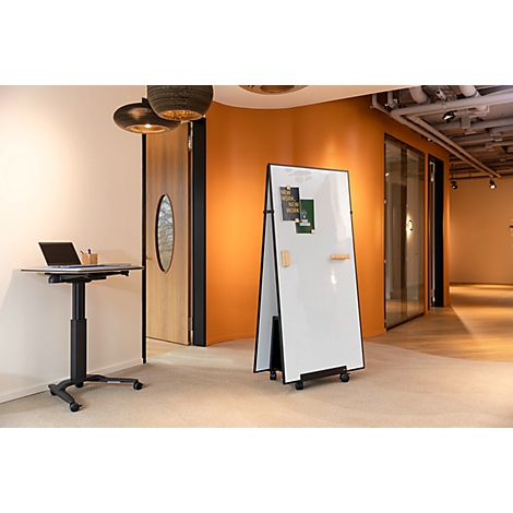 Ein mobiles Whiteboard steht in einem modernen Büro vor einer orangefarbenen Wand neben einem höhenverstellbaren Tisch.