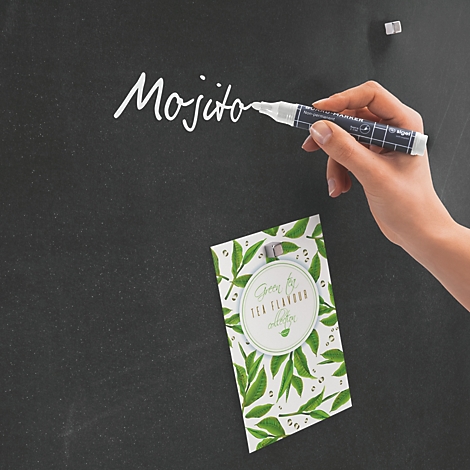 Hand schreibt Mojito mit weißem Stift auf einer schwarzen Tafel. Darunter eine Karte mit grünen Blättern.