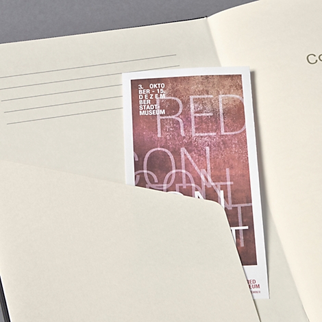 Ein Notizbuch mit einem eingelegten Flyer. Auf dem Flyer steht in Großbuchstaben "RED ON CONT" und Details zu einer Ausstellung.