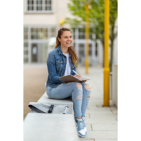 Fröhliche Frau in Jeansjacke sitzt auf einer Mauer und schreibt in ein Notizbuch. Im Hintergrund Gebäude und Bäume.