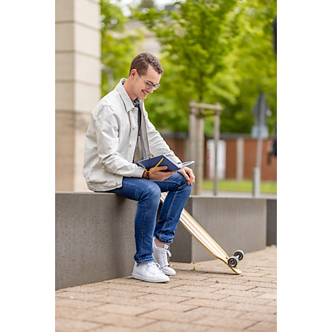 Ein junger Mann mit Brille sitzt draußen und liest in einem Notizbuch. Neben ihm liegt ein Longboard.