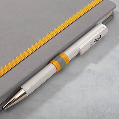 Ein weißer Stift steckt in einem grauen Notizbuch. Der Stift hat einen gelb-grauen Ring und das Logo "sigel".