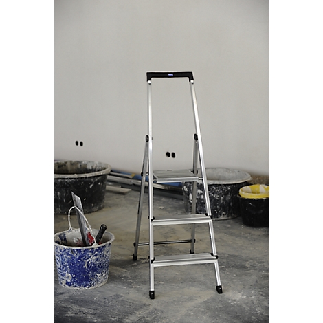 Silver aluminum ladder in front of a white wall, next to buckets and tools. Background: construction site.
