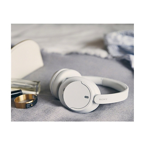 Weiße Kopfhörer von Sony, liegen neben Schmuck und einem Kosmetikartikel auf einer grauen Decke.