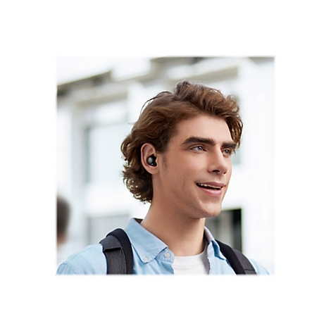 Junger Mann mit In-Ear-Kopfhörern lächelt in die Ferne. Er trägt ein hellblaues Hemd und einen Rucksack.