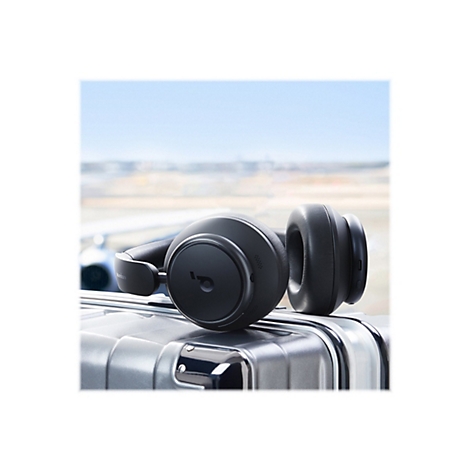 Schwarzer KopfhÃ¶rer liegt auf einem silbernen Koffer. Im Hintergrund ist ein verschwommener Himmel und ein Flugzeug zu sehen.