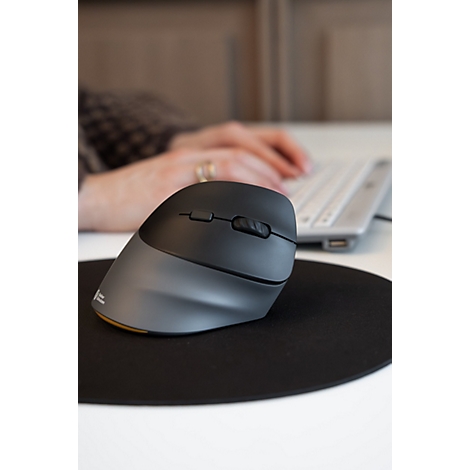 Une souris verticale ergonomique gris foncé sur un tapis de souris noir devant une personne travaillant sur un ordinateur.
