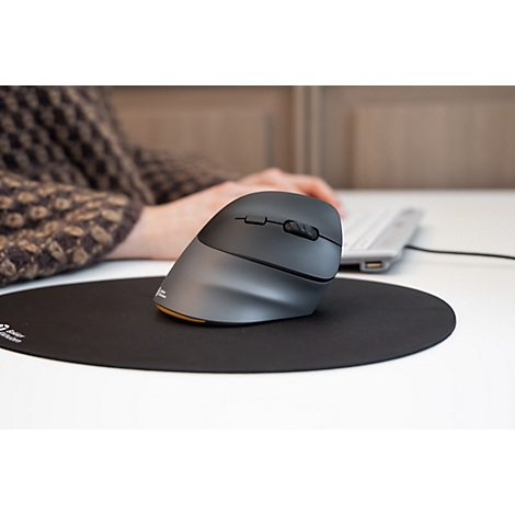 Une souris verticale grise et ergonomique est posée sur un tapis de souris rond et noir, avec une personne tapant au clavier.