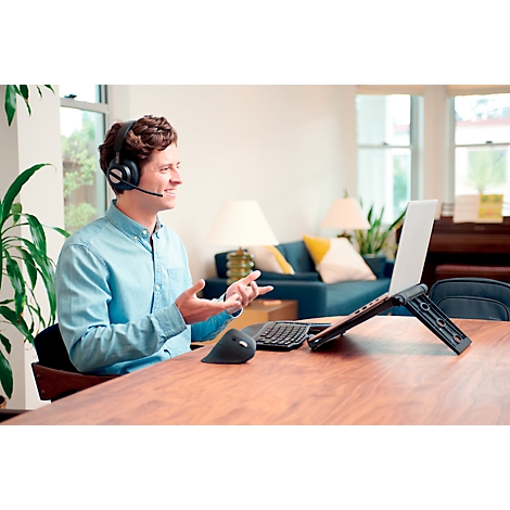 Un homme avec un casque parle sur un ordinateur portable. Il est assis à une table en bois avec une souris et un clavier. Canapé et fenêtre en arrière-plan.