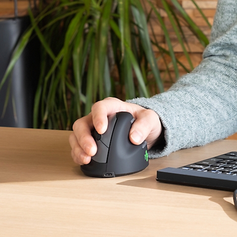 Gros plan sur une main utilisant une souris ergonomique. Le bras de l'utilisateur repose sur la table. Une plante est visible en arrière-plan.