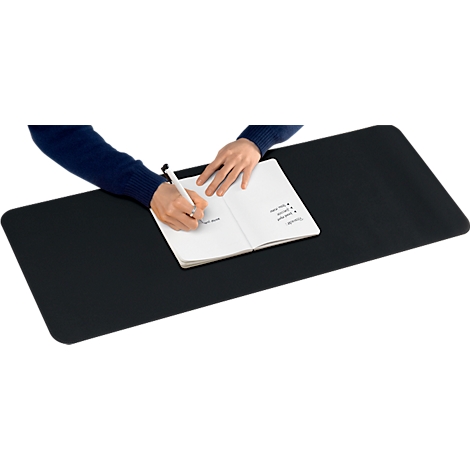 Une personne écrit dans un carnet sur un tapis de bureau noir. Le stylo est dans la main, le carnet est ouvert.