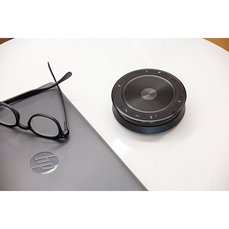 Un haut-parleur noir est à côté d'un ordinateur portable et de lunettes sur une table blanche.