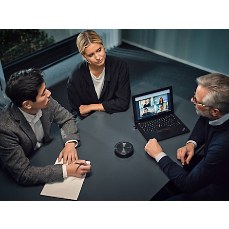 Trois personnes sont assises à une table utilisée pour une vidéoconférence. Une vidéoconférence avec plusieurs participants peut être vue sur l'ordinateur portable.