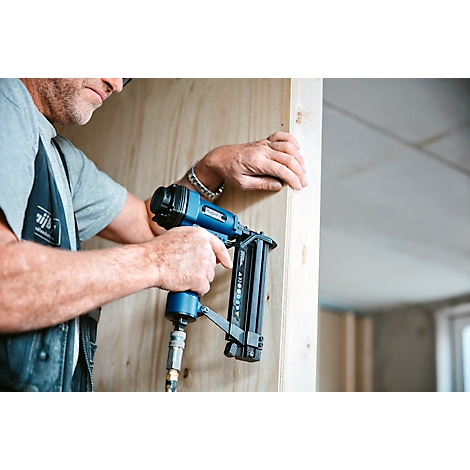 Een timmerman gebruikt een blauwe pneumatische tacker. Hij bevestigt hout aan een muur. Grijze achtergrond.