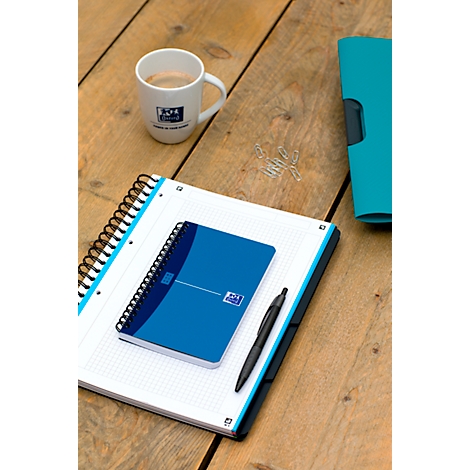 Een blauw notitieboek, een kleiner notitieboek, pen, koffiekop en paperclips op hout. De achtergrond is licht wazig.