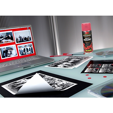 A workstation with a laptop, photos, adhesive spray, and CDs. The laptop displays images on a red interface.