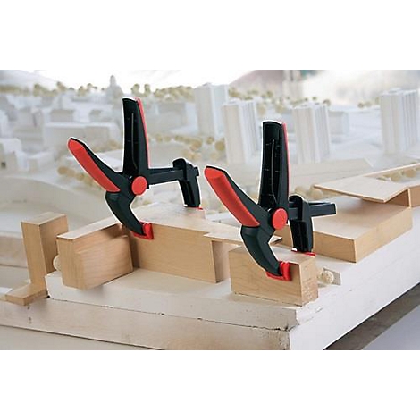 Two black and red clamps secure pieces of wood on a white model base. A city model is visible in the background.