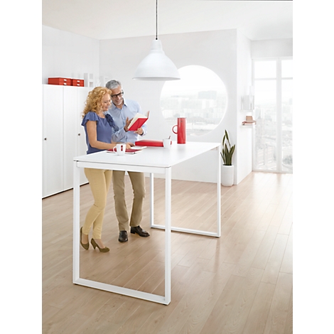 Two people stand at a white table, looking at a red book. Bright interior with a large window.