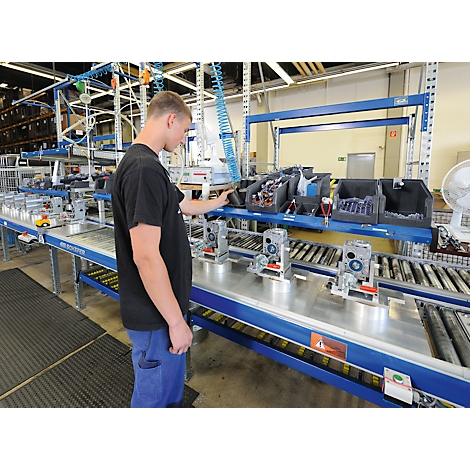 Un homme en t-shirt noir se tient près d'une chaîne de montage dans une usine et contrôle des machines.
