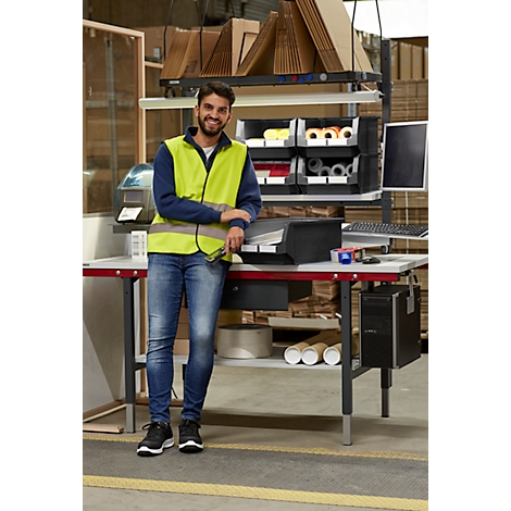 Un homme sourit près d'une table de travail dans un entrepôt, portant un gilet de sécurité. En arrière-plan, des étagères et un ordinateur.