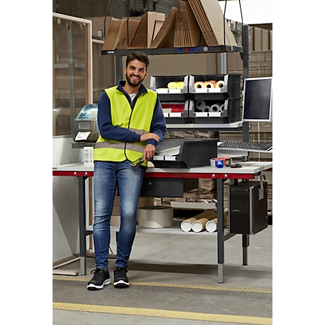 Un homme en gilet jaune s'appuie sur une table dans un entrepôt, souriant à la caméra. Des étagères et des ordinateurs sont derrière lui.
