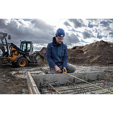 Een bouwvakker knielt bij de fundering van een bouwplaats en meet met een geel meetlint. Op de achtergrond een graafmachine en donkere wolken.