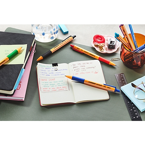 Nahaufnahme eines Schreibtischs mit Schreibwaren. Geöffnete Notizbücher mit handschriftlichen Notizen. Mehrere Stifte in verschiedenen Farben liegen herum.