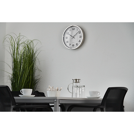 A table in a meeting room. Coffee cups, a glass jug and a vase with tall grass are on the table. A clock hangs on the wall.