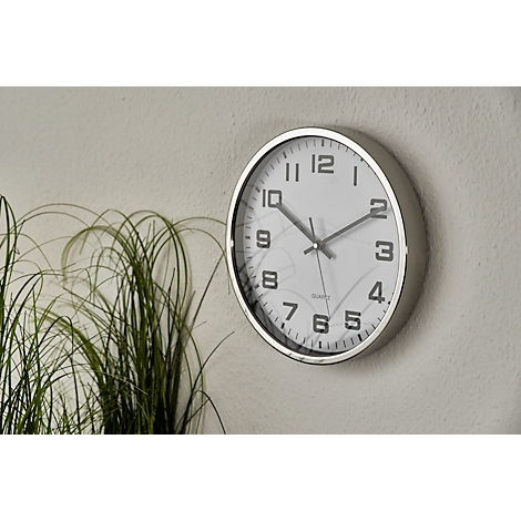 A round silver wall clock with black markings hangs on a light grey wall. Green plants are visible behind it.