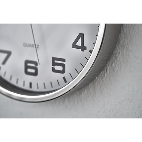 Close-up of a clock: dial with numbers 4, 5, 6 and 7, and a silver case, attached to a textured gray wall.