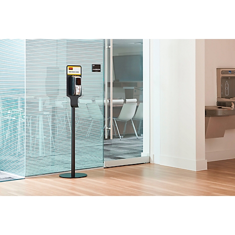 Een zwarte handdesinfectiemiddeldispenser staat in een lichte kamer. Achter glazen wanden is een vergaderzaal te zien.