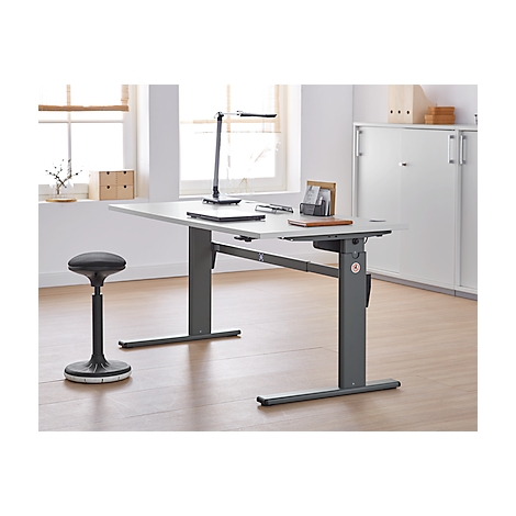 An adjustable height desk, a lamp, and a stool in an office. A window and a cabinet in the background.