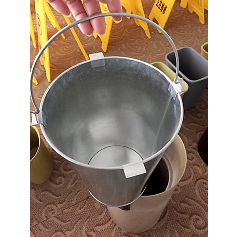 A person holds an empty metal bucket with a handle over a waste bin. In the background, yellow signs warning of slippery surfaces.