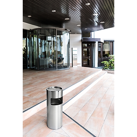 Stainless steel ashtray in front of an entrance hall. Revolving door, stairs and shiny ceiling in the background.