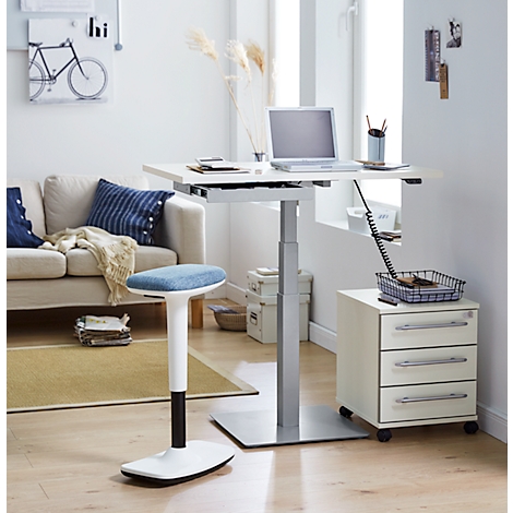 An adjustable height desk with a laptop, standing stool, drawer cabinet, and sofa in a bright room.