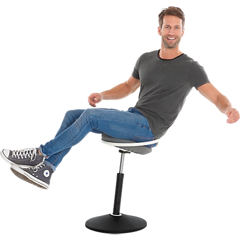 A man sits on a height-adjustable chair. He is smiling and has his arms outstretched. He is wearing jeans, a grey T-shirt and Chucks.