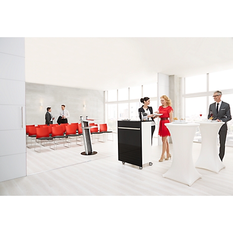Several people are standing at high tables in a conference room. Red chairs are ready. Conference technology in the background.